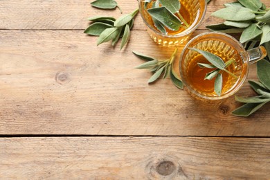 Cups of aromatic sage tea and fresh leaves on wooden table, flat lay. Space for text