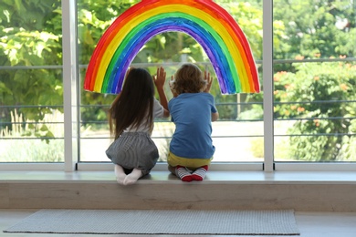 Photo of Little children sitting near rainbow painting on window indoors. Stay at home concept