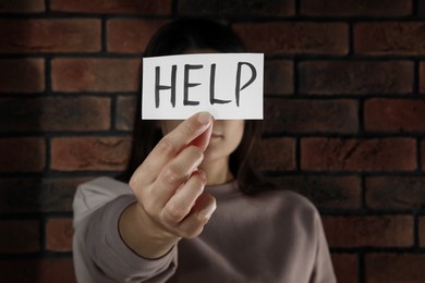 Photo of Domestic violence concept. Woman holding paper with written word Help near brick wall, focus on hand
