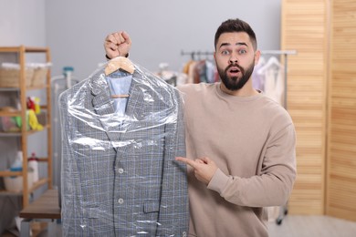 Photo of Dry-cleaning service. Shocked man holding hanger with jacket in plastic bag indoors