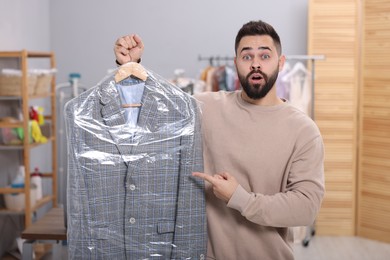 Dry-cleaning service. Shocked man holding hanger with jacket in plastic bag indoors