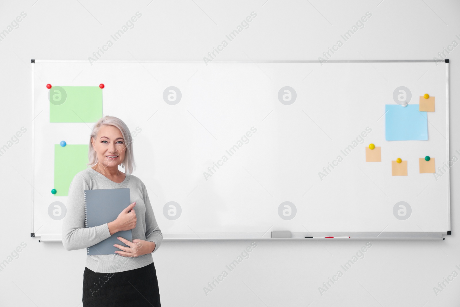 Photo of Mature teacher with notebook near whiteboard in classroom. Space for text