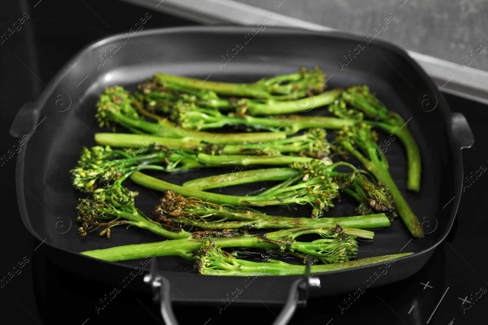 Photo of Grill pan with tasty cooked broccolini on cooktop, closeup