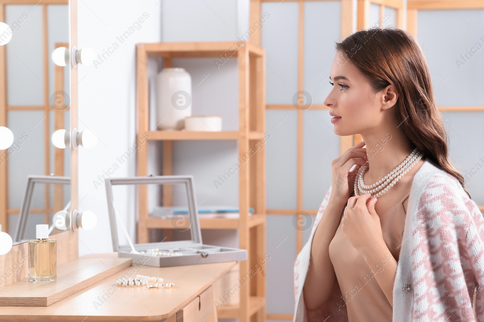 Photo of Young woman trying on elegant pearl necklace at dressing table indoors