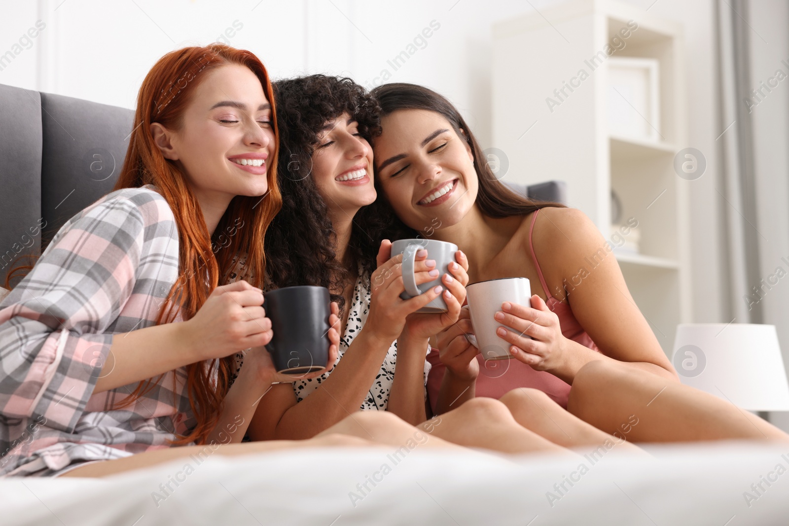 Photo of Happy young friends with cups of drink on bed at home