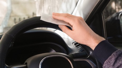 Man cleaning steering wheel with wet wipe in car, closeup. Protective measures
