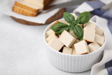 Photo of Bowl of smoked tofu cubes with basil on white table, closeup. Space for text