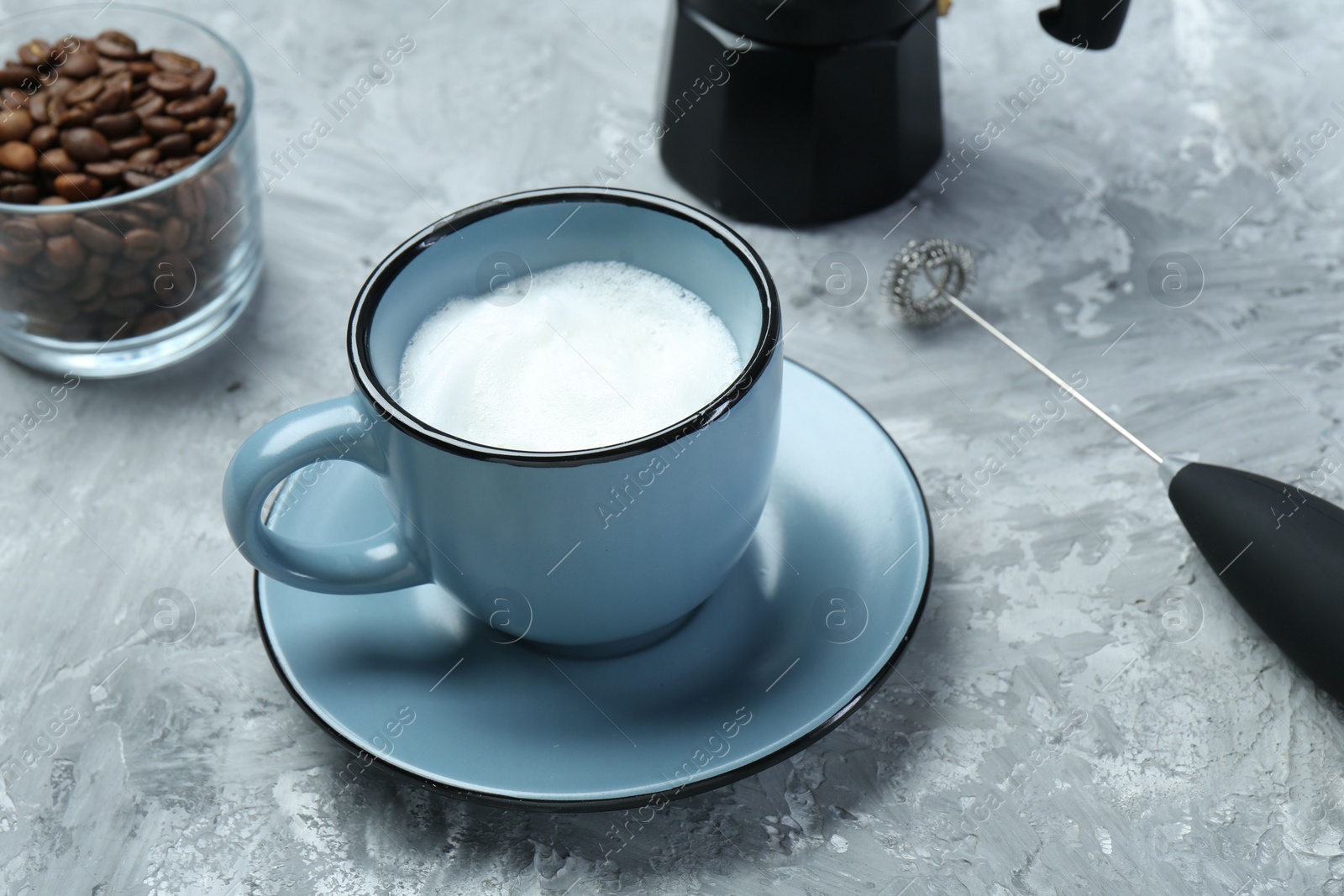 Photo of Mini mixer (milk frother), whipped milk in cup and coffee beans on grey textured table