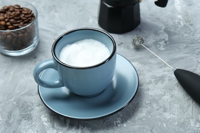 Photo of Mini mixer (milk frother), whipped milk in cup and coffee beans on grey textured table