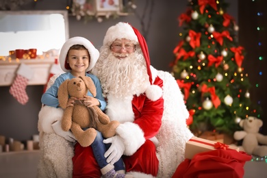 Photo of Little boy with toy bunny sitting on authentic Santa Claus' lap indoors