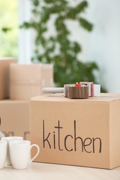 Photo of Cardboard box, cups and adhesive tape dispenser on table indoors. Moving day