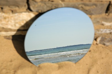 Photo of Round mirror reflecting sea on sand near stone wall outdoors, closeup