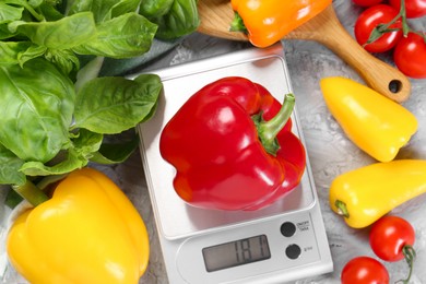 Kitchen scale with bell pepper among basil and tomatoes on grey textured table, flat lay