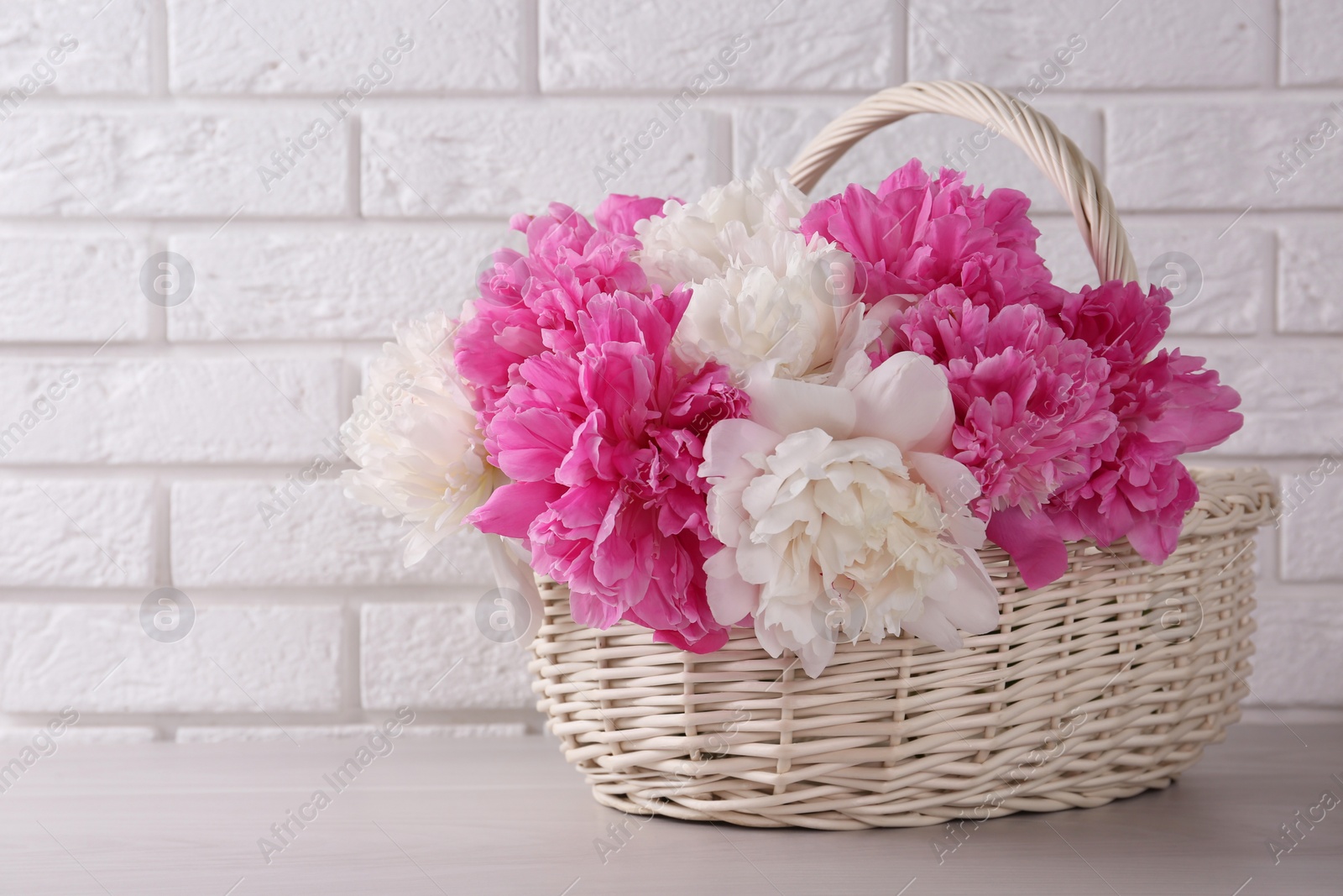 Photo of Beautiful peonies in wicker basket on white table near brick wall