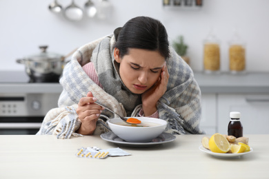 Sick young woman eating soup to cure flu at table in kitchen