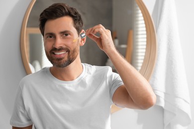 Smiling man applying cosmetic serum onto his face in bathroom. Space for text