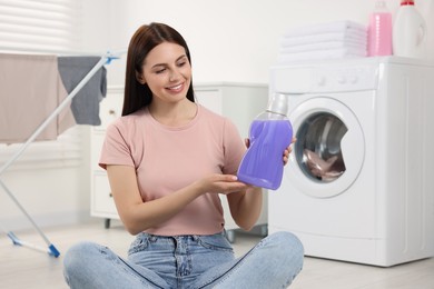 Photo of Woman sitting near washing machine and showing fabric softener in bathroom