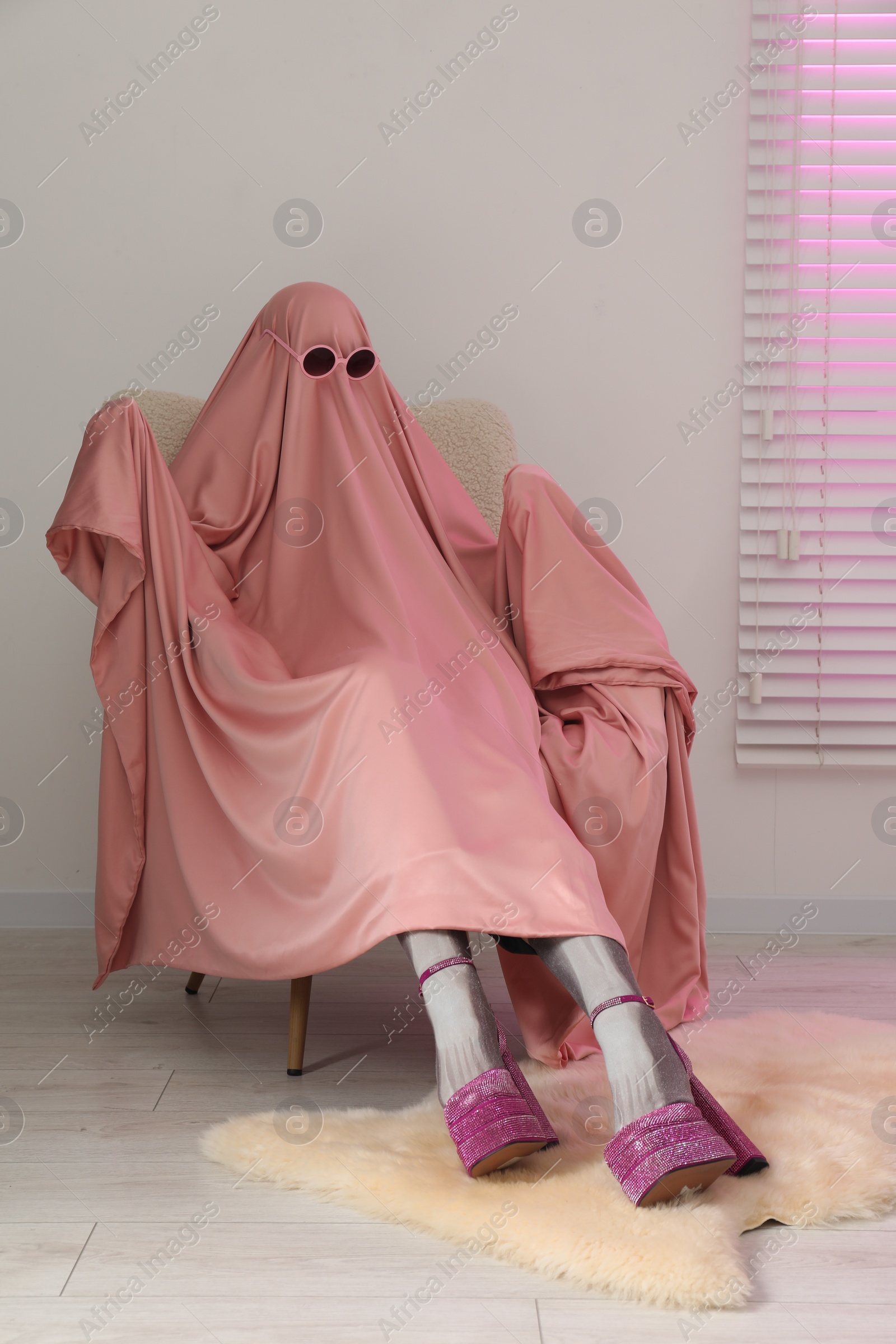 Photo of Glamorous ghost. Woman in pink sheet and high heel shoes on armchair indoors