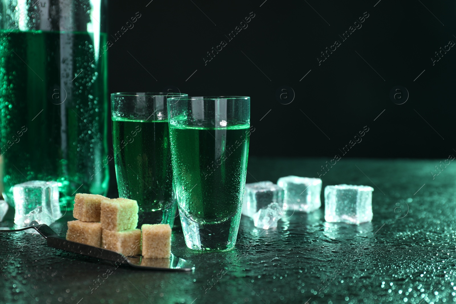 Photo of Absinthe in shot glasses, spoon, brown sugar and ice cubes on gray table against dark background, space for text. Alcoholic drink