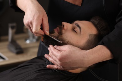 Photo of Professional barber shaving client's beard with blade in barbershop, closeup