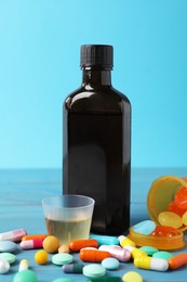 Photo of Bottle of syrup, measuring cup and pills on light blue table. Cold medicine