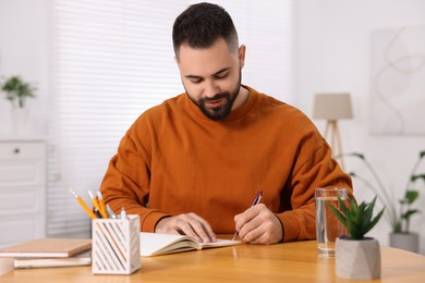 Photo of Young man writing in notebook at wooden table indoors