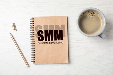 Image of Notebook with text SMM (Social media marketing, cup of coffee and pen on white wooden background, flat lay