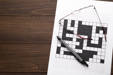 Photo of Blank crossword, glasses and pen on wooden table, top view. Space for text