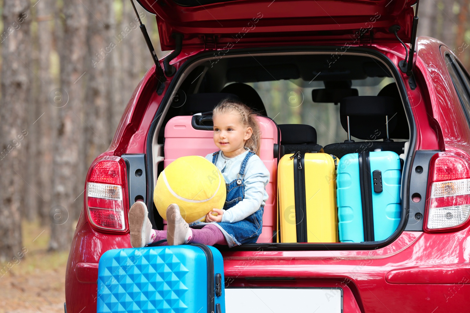 Photo of Cute little girl sitting in car trunk loaded with suitcases on forest road