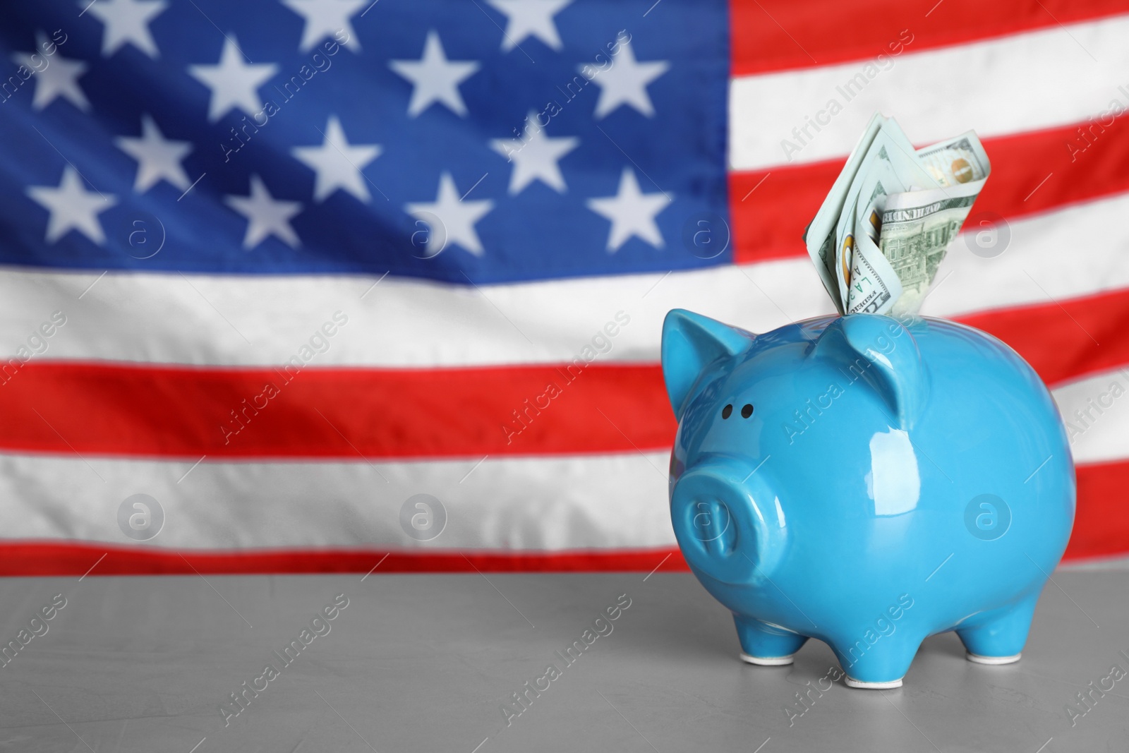 Photo of Blue piggy bank with banknotes on light grey table against national USA flag. Space for text