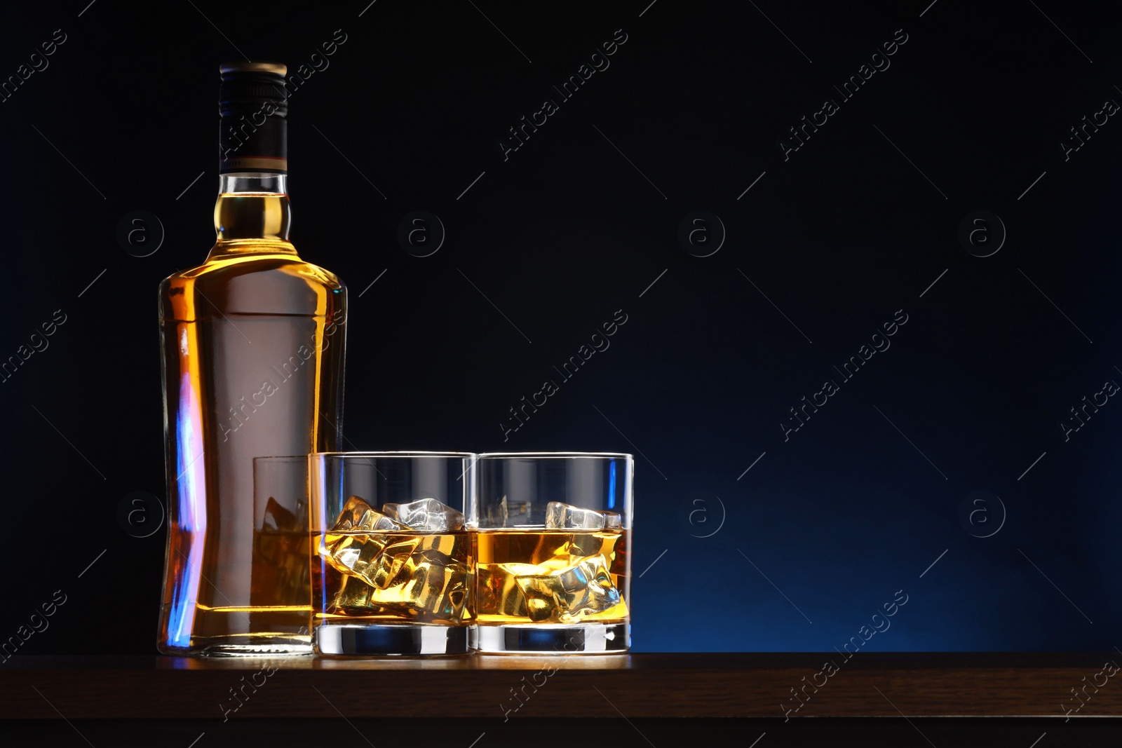 Photo of Whiskey with ice cubes in glasses and bottle on table against dark background, space for text