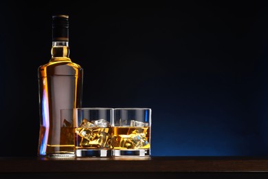 Whiskey with ice cubes in glasses and bottle on table against dark background, space for text