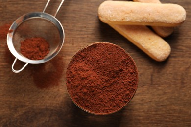 Delicious tiramisu in glass, biscuits and sieve with cocoa powder on wooden table, flat lay