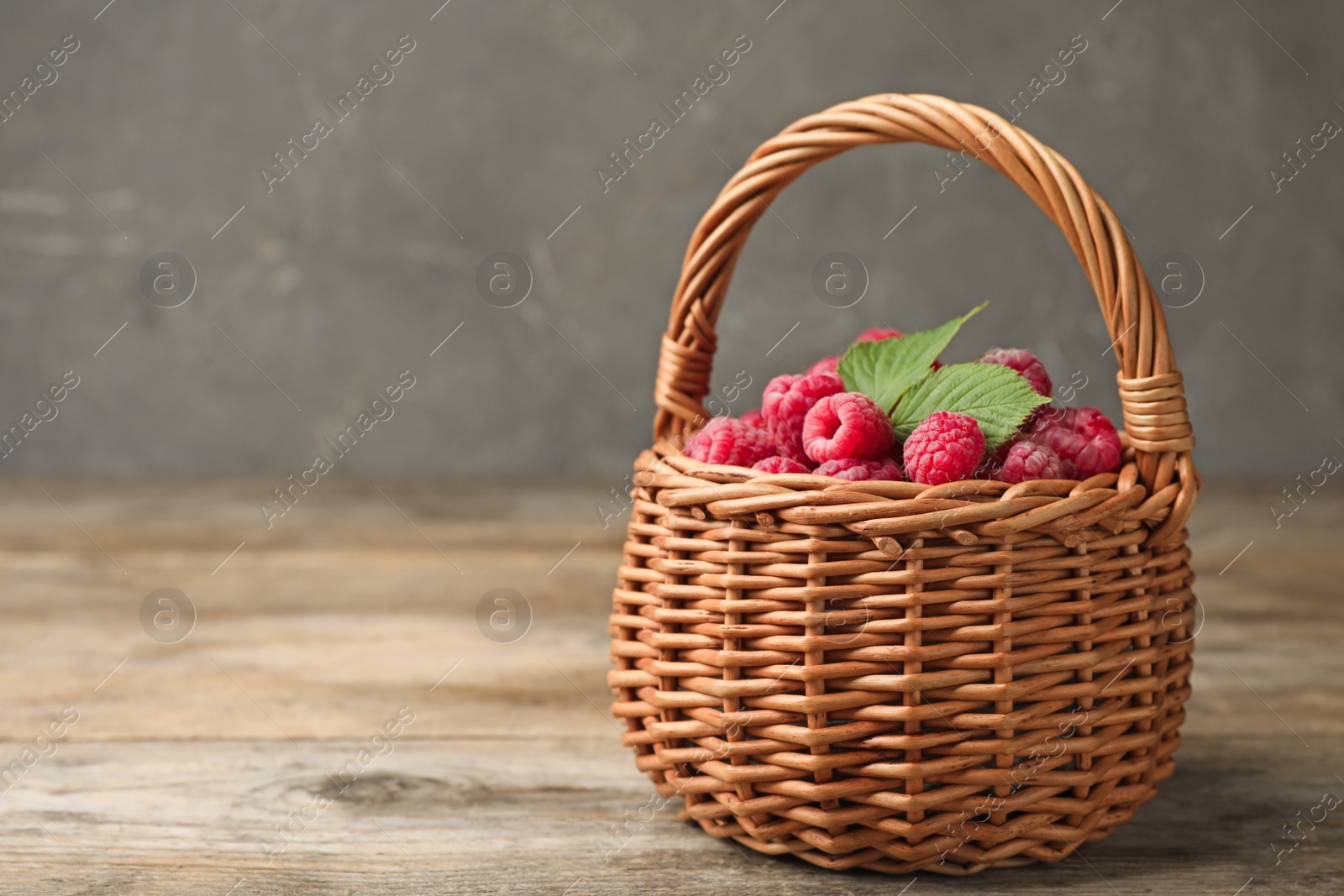 Photo of Basket of delicious fresh ripe raspberries with leaves on wooden table against grey background, space for text