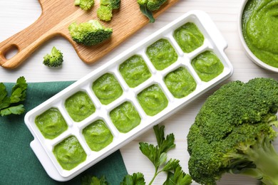 Photo of Broccoli puree in ice cube tray and ingredients on white wooden table, flat lay. Ready for freezing