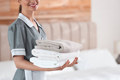 Young chambermaid holding stack of fresh towels in bedroom, closeup