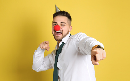 Emotional young man with party cap and clown nose on yellow background. April fool's day