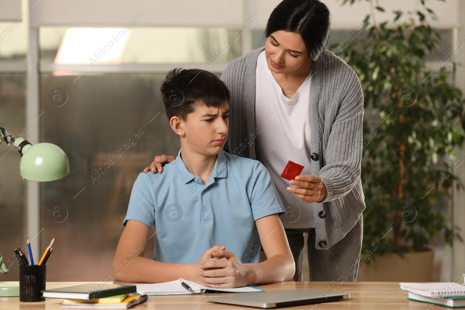 Photo of Mother giving condom to her teenage son while he doing homework at home. Sex education concept