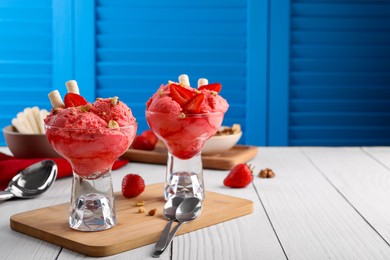 Photo of Delicious scoops of strawberry ice cream with wafer sticks and nuts in glass dessert bowls served on white wooden table. Space for text