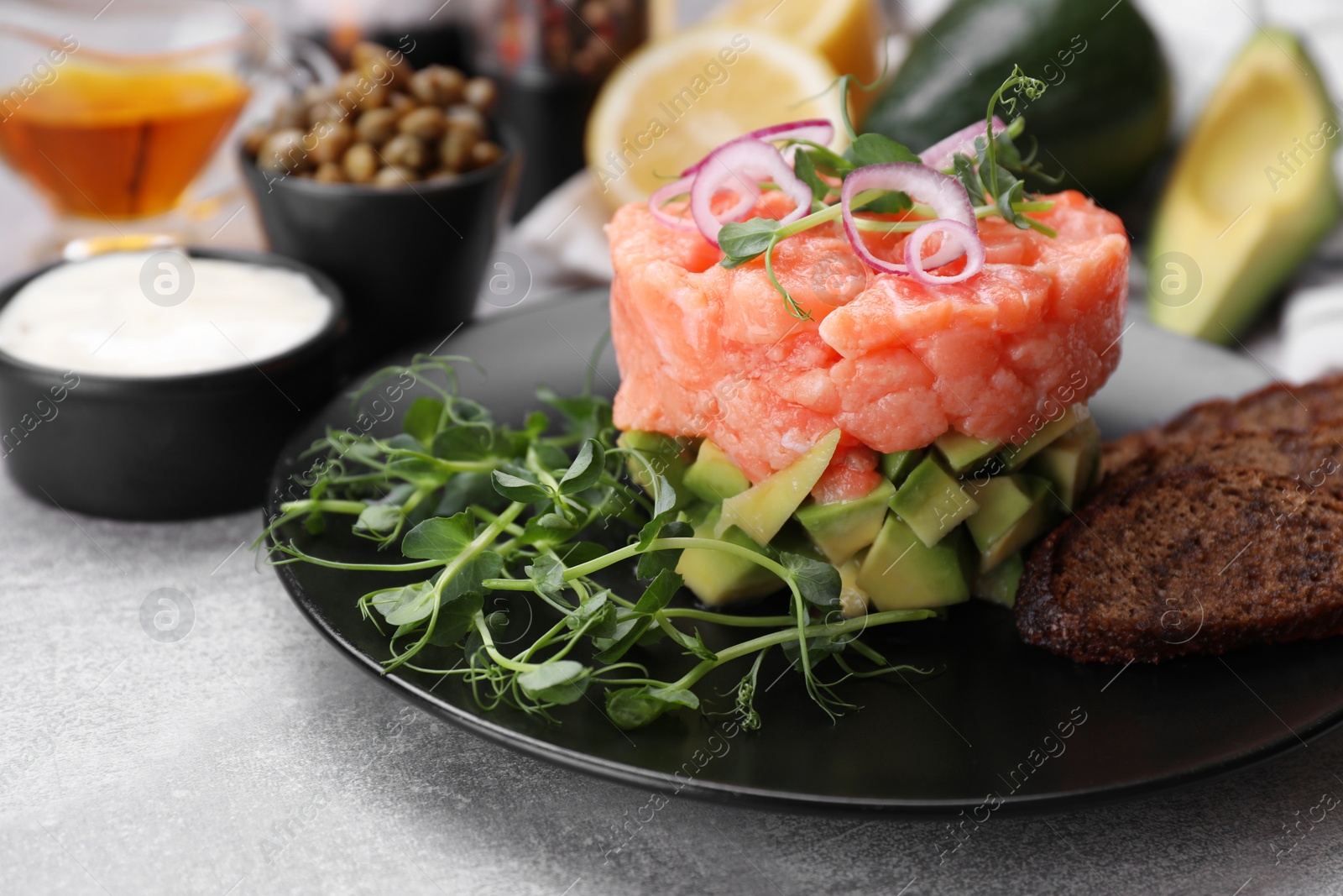 Photo of Tasty salmon tartare with avocado, microgreens and croutons on grey table, closeup