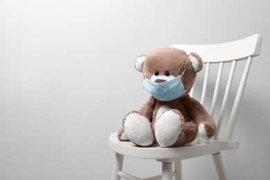 Photo of Cute teddy bear in medical mask on chair near light wall, space for text