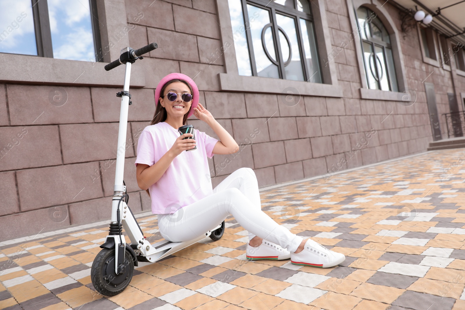 Photo of Young woman with cup of coffee sitting on electric kick scooter outdoors