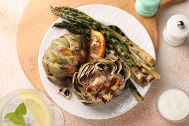 Photo of Tasty grilled artichokes and asparagus served on beige textured table, flat lay