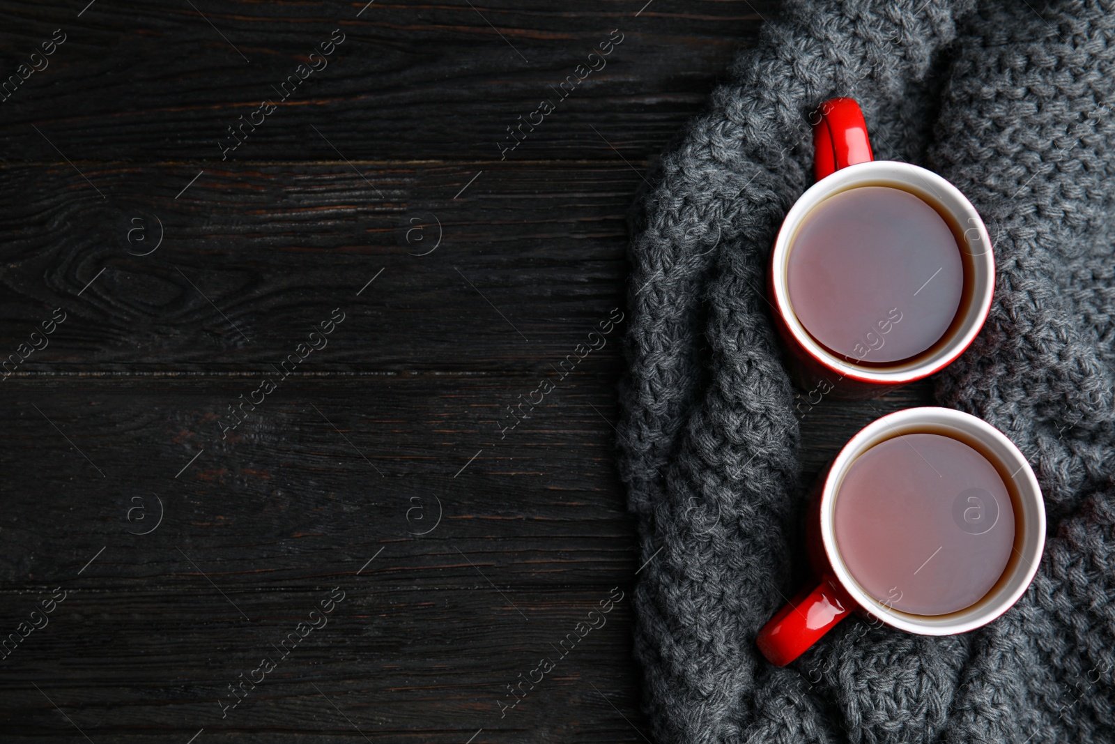 Photo of Flat lay composition cups of hot tea and knitted scarf on black wooden background, space for text. Winter drink