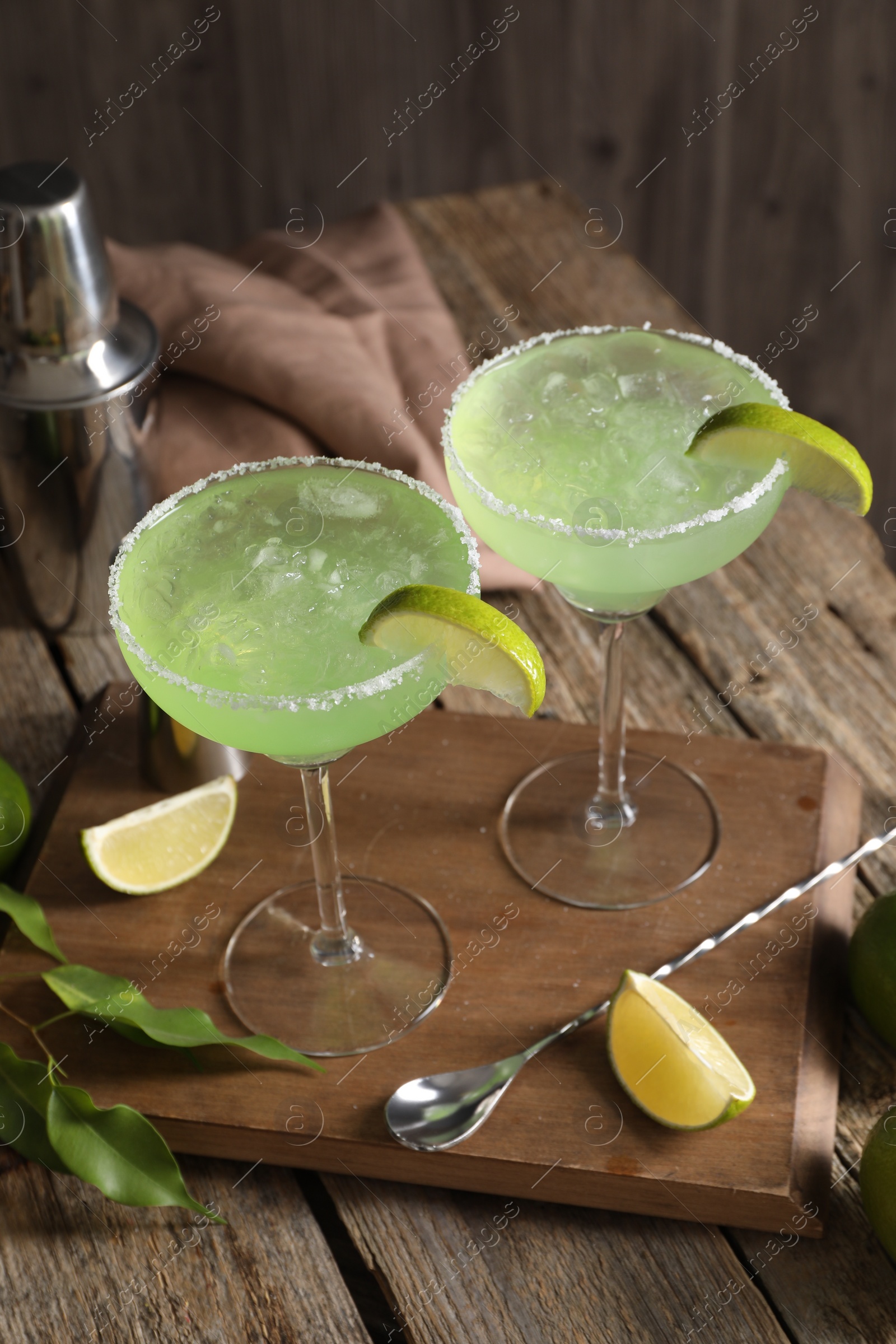 Photo of Delicious Margarita cocktail in glasses, limes, bar spoon and shaker on wooden table