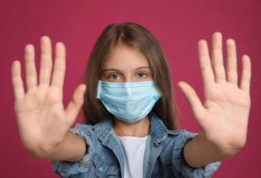 Photo of Little girl in protective mask showing stop gesture on crimson background. Prevent spreading of coronavirus