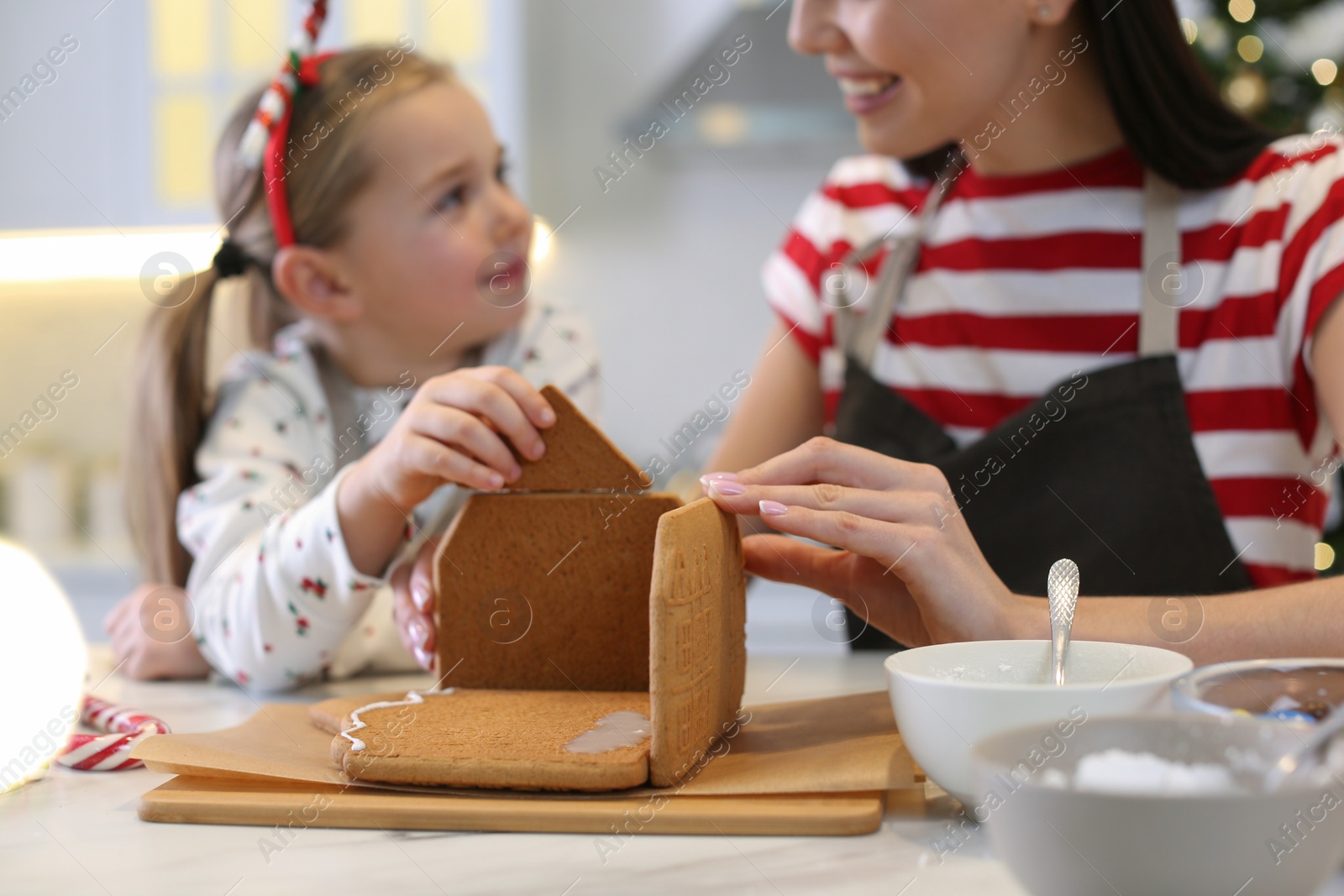 Photo of Mother and daughter making gingerbread house at table indoors, closeup