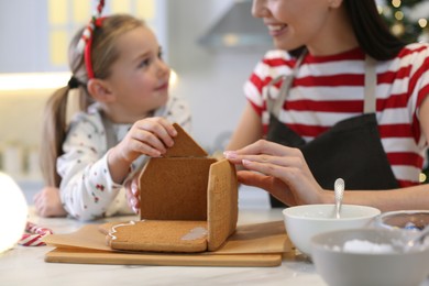 Photo of Mother and daughter making gingerbread house at table indoors, closeup