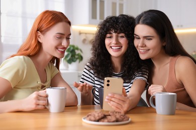 Photo of Happy young friends with smartphone spending time together at table in kitchen