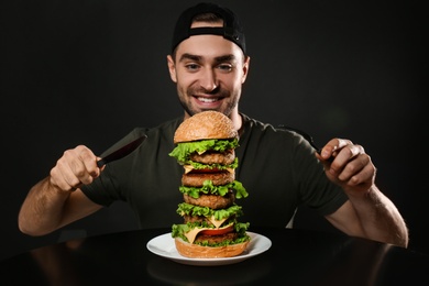 Young man with cutlery eating huge burger on black background